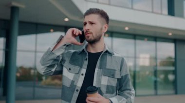 Genç sakallı bir adam telefonda konuşuyor. Şehir ortamında endişeli görünüyor. Endişeli Sakallı Genç Adam Şehir Ayarlarında Akıllı Telefonla Konuşuyor