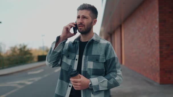 街の屋外で 若いひげ付きの男が電話で話し 深刻で不満を表しています スマートフォン経由で深刻な会話に従事したヒスパニック男性 — ストック動画