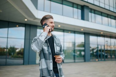 Modayı takip eden bir adamın elinde kahveyle akıllı telefonuyla konuştuğu anı kutluyoruz. Dijital Diyalog Şehir Çevresindeki Akıllı Telefondaki Gülümseyen Erkek