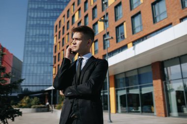 Şehir siluetinin arka planında, takım elbiseli genç bir adam akıllı telefonuyla bir konuşmaya aktif olarak katılıyor, beyaz yakalı bir ortamda modern profesyoneli temsil ediyor..