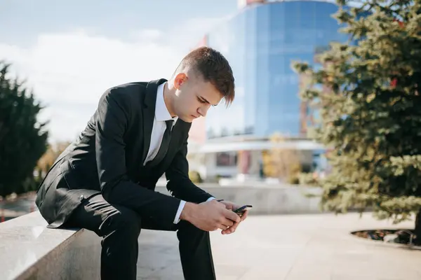 Within the vibrant urban business district, a young and dedicated professional sits outdoors, deeply engaged with his smartphone. His business suit sets the tone for his commitment to success.