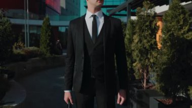 Şık giyinmiş ve güneş gözlüklü bir erkek işadamı, şehir manzarasında kol saatini dikkatle izliyor ve zaman yönetimini vurguluyor..