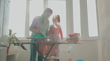 Genç Anne ve Kız Ev İşi İçin Ev ve Aile Sıcaklığını Kucaklıyorlar. Evde Anne ve Çocuk Ütüleme Kıyafetleri Birlikte