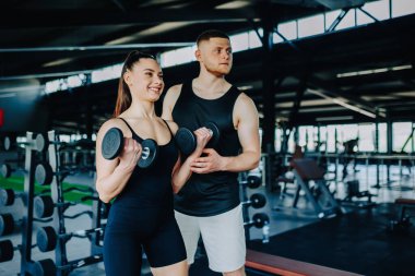Erkek bir kişisel eğitmenin rehberliğiyle, bir kadın spor salonunda başarılı bir şekilde dambılı kaldırır. Jimnastik Salonu Oturumu Erkek Eğitmeni Aptallar Egzersizinde Kadına Yardım Ediyor