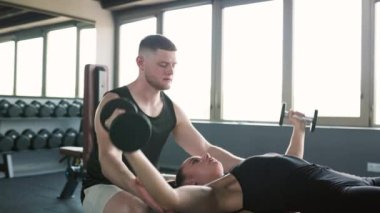 Kendini adamış bir erkek antrenör, spor salonunda bir kadına dambılı kaldırmasında yardımcı olur. Spor Salonu Erkek Antrenörü Aptal Kaldıran Kadın Asistanı