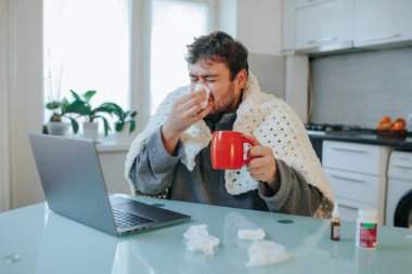 Canlı bir görüntüde, sakallı genç bir adam, mutfak sandalyesine sarılı bir yorgana sarılmış, griple mücadele ediyor. Titremesi ve burnunu silmesi mevsimsel hastalıkları yakalar..