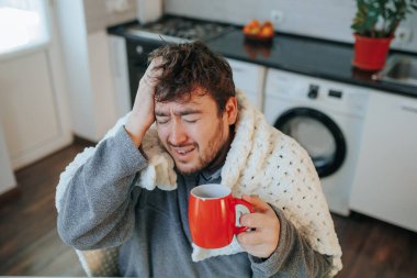 Stresli bir adam, sakallı ve yorgana sarılı, baş ağrısı ve evde grip geçiriyor. Mutfak sandalyesindeki bu nüanslı tasvir mevsimsel hastalıkların zorluklarını yakalar..