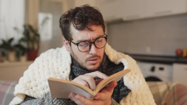一个人舒适地躺在被褥下 在客厅里翻阅着一本书 眼镜增加了他的智力氛围 书虫窝无暖气公寓 — 图库视频影像
