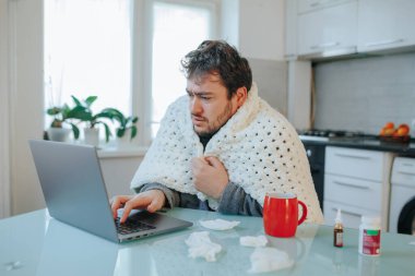 Frosty Workday Grit Genç sakallı bir adam sıcak bir yorganın altında griple yüzleşiyor, soğuk bir evde dizüstü bilgisayarına odaklanıyor..