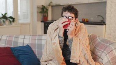 Evde griple savaşan bir adamın resmi. Gözlüklü, sıcak bir içeceği yudumlarken, oturma odasında yorganla kaplanmış, hastalığın ve soğuk havanın kesiştiği yeri gösteriyor..