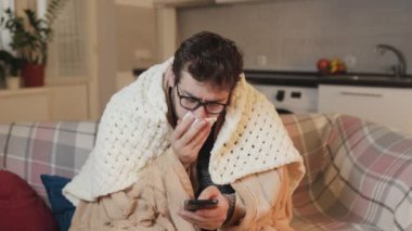 Gözlüklü Grip Genç Adam 'ın Ortasında Dijital Bağlantı Smartphone' da, Evde Hastalıkla Mücadele. Oturma odasında bir yorganla titreyerek ve üzerini örterek Soğuk Gerçekliği iletiyordu.