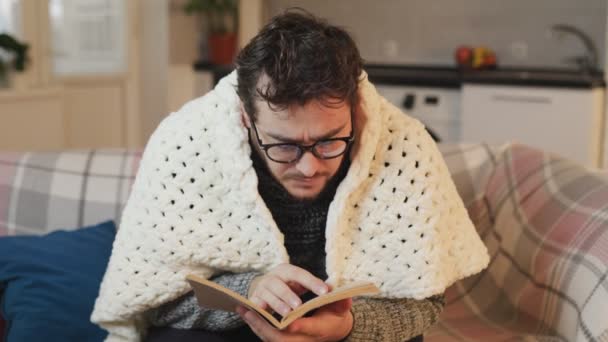 寒い冬の読書 メガネの男性リーダーは 彼の無熱のリビングルームで快適さを見つける羽毛布団の下で本を楽しんでいます ホーム コンフォート 無熱リビングルーム 読書セッション — ストック動画