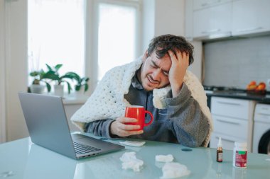 Donmuş Çalışma Alanı Mavisi Sakallı adam, dağınık saçlı, grip olmuş, yorganın altında, dizüstü bilgisayarın yanında, soğuk bir günde evde savaşan.