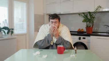 Yorgana sarılmış sakallı, genç, beyaz bir adam griple baş ederken stres ve acı çekiyor. Evde bir mutfak sandalyesine oturmuş, hapşırıyor ya da öksürüyor.