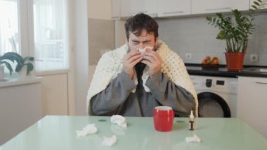 Bu samimi görüntüde, yorganın içine sarılı genç bir adam mutfak sandalyesindeki gripte geziniyor. Titremesi ve burnunu silmesi evdeki mevsimsel hastalıkların sorunlarını gösteriyor..