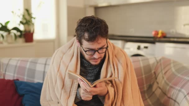 メガネをかぶった男が 羽毛布団に包まれて 彼の無熱のリビングルームで解決策を見つけた デュヴェット デイズ コールド ウェザー リーディング リビング — ストック動画