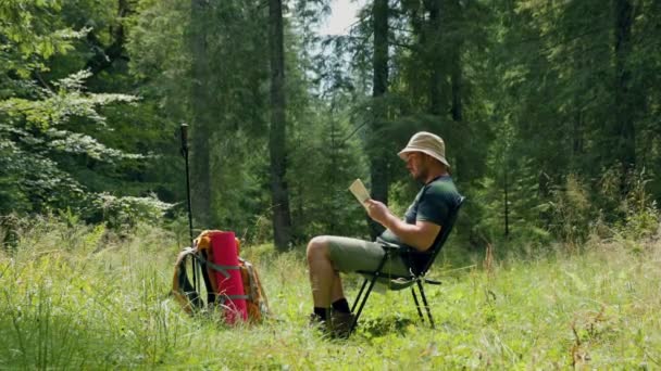 和一个年轻的旅行者一起在树林中进行文学旅行 在那里 阅读的行为在大自然的心脏变成了一种宁静的仪式 徒步旅行者坐在椅子上 手里拿着一本书 — 图库视频影像