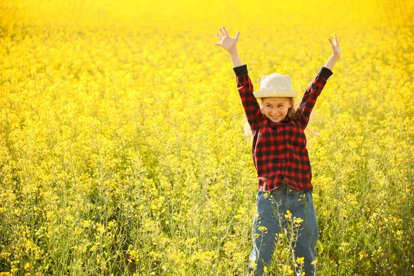 front view of little kid girl with hat looking at camera raise your hands up in flowering rapeseed land. happy little girl jumps up