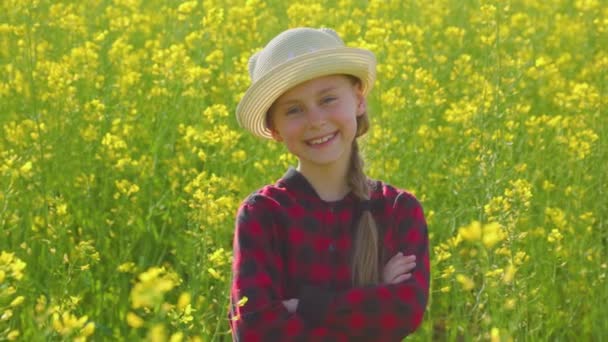 美丽的笑容可亲的小女孩 头戴一顶黄色的花油菜花帽 抱着她的胳膊 看着相机 小孩笑了 — 图库视频影像