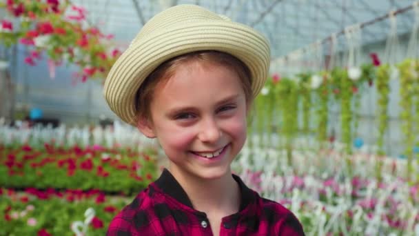 花の温室で頭に帽子をかぶった笑顔の女の子の肖像画 彼女はそれらを世話する植物に満足しています カメラを見る — ストック動画