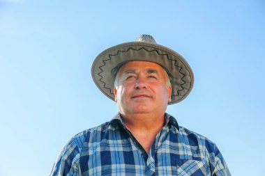 Güneşli bir yaz günü kırsal kesimde tarım çiftliğinde dikilen şapkalı yaşlı, mutlu erkek işçi kırsal kesimde mutlu ve olgun bir çiftçi.