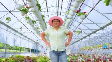 Woman aged florist stands in greenhouse with flowers, she smiles and puts her hands on her hips. Dressed in a hat, shirt and jeans. The greenhouse is light and there are many houseplants. Copy space.