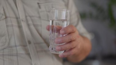 Bu görüntü, yaşlı bir adamın ellerini, dudaklarına bir bardak suyu nazikçe kaldırırken, özbakım ve hidratasyon anını somutlaştıran yakın çekim görüntüsü sunuyor.