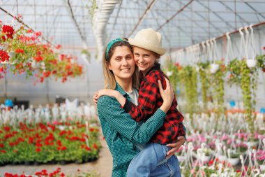 Kırmızı kare gömlekli ve şapkalı neşeli bir kız çocuğunun annesinin ellerinde oturması. Mutlu güzel kız ve anne ailesi kameraya bakıp serada poz veriyor. Bahçıvan ailesi, kopyalama alanı