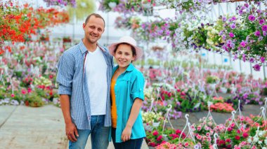 Young happy man and woman rejoice at their success while standing in a greenhouse with flowers. Big glasshouse and next to each other. Young people are dressed in casual style.