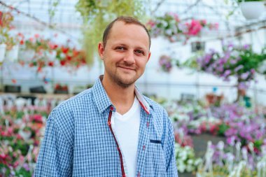 A young handsome farmer stands in a greenhouse and looking at camera. The head is slightly tilted to the left. The man smiles a calm smile. The agronomist is dressed in a plaid shirt. Copy space