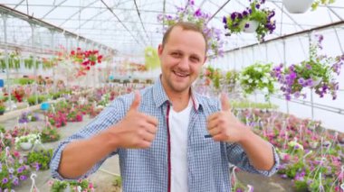 A very happy lucky man stands in a greenhouse and shows thump up with both hands. The businessman laughs heartily and shows how successful everything is. Dressed in a plaid shirt.