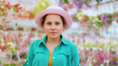 Young nice agronomist stands in a greenhouse and looks into the camera. The woman turned her head to the right. Girl smiles calmly. The agronomist is dressed in a blue shirt and has a hat on had.