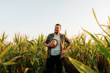 Tarlada yüzünde gülümseme olan düşünceli bir çiftçi, şapkasını çıkardı, yorgun, önden, mısır tarlasının arka planında genç bir adam, bir eli kalçasında, açık gökyüzü. Boşluğu kopyala.