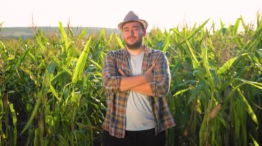 Şapkalı genç çiftçi mısır tarlasında mutlu bir şekilde duruyor, başını çeviriyor ve kameraya gülümsüyor, kameraya bakıyor. Güneş tepemizde ışıldıyor,