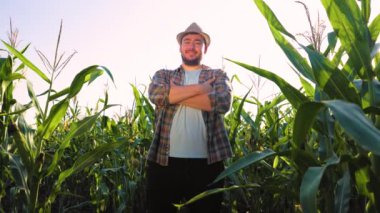 Bottom view of a young male farmer in a hat is standing in a corn field looking forward with his arms crossed. The end of the working day, the man is happy from the work done. Beautiful sunset.