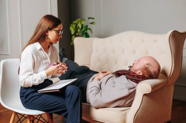 Elderly male client of psychotherapy woman relaxed lying on couch in office, girl says. Psychological session, consultation of psychologist, comfortable posture conducive to confidential conversation. clipart