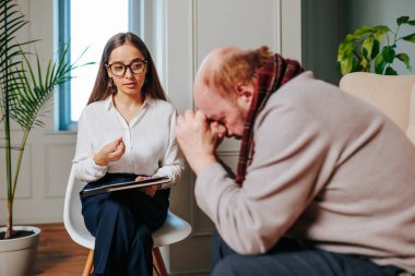 Genç kadın psikolog müşterisine, elleriyle başını destekleyen yaşlı bir adama bir şey söylüyor. Yaşlıların sorunları, psikologların yardımı, psikoterapist, yaşlanma psikolojisi.
