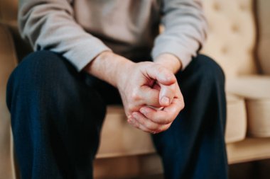 Tanımlanamayan yaşlı bir adamın seçici odak noktası. İçeride bir adam kanepede oturuyor. Elleri birbirine bağlıdır. Arka plan bulanık. Ön görünüm.