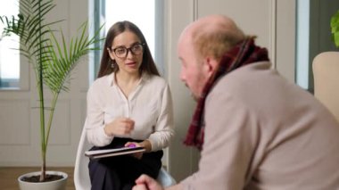 Yaşlı bir adam, elleri ile başını dik olarak ofiste oturur, kadın psikolog dikkatle dinler. Geniş, parlak psikoterapi ofisi, psikolojik yardım, yaşlılık, bunama hastalıkları..