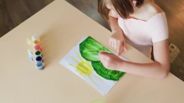 Üst tabaka tanınmayan çocuk evde masada oturuyor ve bir resim çiziyor. Ev eğlencesi. Güzel çiziyor. Renkli resim. Kız evde kağıt, su rengi ve sanat fırçasıyla resim yapmaktan zevk alıyor..