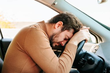Genç bir araba sürücüsü direksiyonda uyuyor, elleri direksiyonda, başı orada yatıyor. Yolda çok fazla zaman geçirmek yorgunluğa yol açar. Fazla mesai yapamazsın..