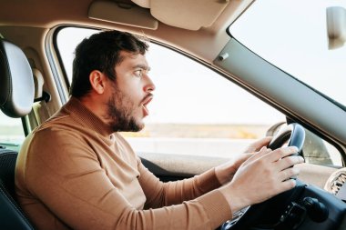 Direksiyonda oturan bir erkek sürücünün yüzünde şaşkınlık, öfke ve öfke ifadesi. Bir adam araba sürerken çok şiddetli duygular ifade eder. Farklı insani duygular..