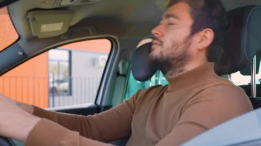 Bir arabanın direksiyonundaki çok üzgün genç bir adam dinlenmek için yatıyordu. Şehir hayatının koşuşturması rahatlamana izin vermiyor. Sakal ve kahverengi kazak..