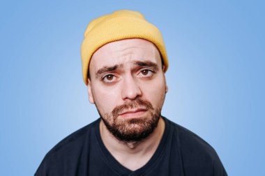 Sarı şapkalı, mağlup bir ifadeyle yorgun, mutsuz bir adamın portresi. Mavi arka planı olan bir stüdyoda oturuyor. Yorgunluk ve duygusal yorgunluk hissi veren bir adam. Boşluğu kopyala