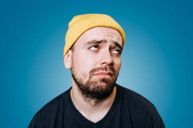 Stüdyo çekimlerinde mavi izole edilmiş arka planda duran yorgun ve uykulu beyaz yakışıklı tuhaf adamın portresi. Sarı şapkalı.
