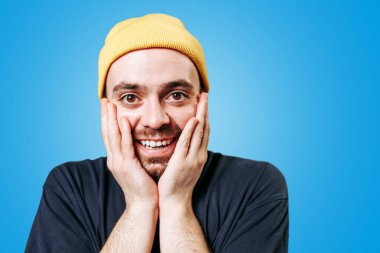 Sarı şapkalı, şaşkın sakallı bir adam, mavi arka planda bir stüdyo resmi, şaşırmış ve şaşırmış bir bakış atıyor..