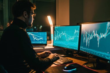 The Home-Based Trader of a man using his computer monitor and calculator at night time as he works on his homework, making trades and analyzing financial data from the comfort of his home office.