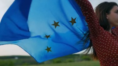 Açık mavi gökyüzünün arka planında, bir kadın Avrupa Birliği bayrağını tutkuyla sallıyor, Avrupa ulusları arasındaki dayanışma ve işbirliği ruhunu somutlaştırıyor..