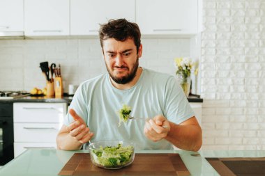 Genç bir adam elinde çatalla sulu yeşil bir salata tutarken tiksinme duygusunu gösterir. Masada bir kase sağlıklı yemek var, ama kimse onu yemek istemiyor. Diyet yapma.