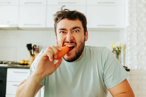 stock image Front view looking at camera a young man makes a grimace and bites raw carrots with anger, does not like vegetables. Does not want to go on a meat free diet. Vegan food spoils the mood. Stop diet.